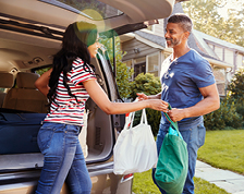 A woman with long hair stands in front of an open SUV trunk and hands full bags over to a man.